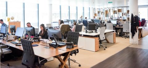 Een open kantoortuin met medewerkers achter bureaus met beeldschermen en telefoons.